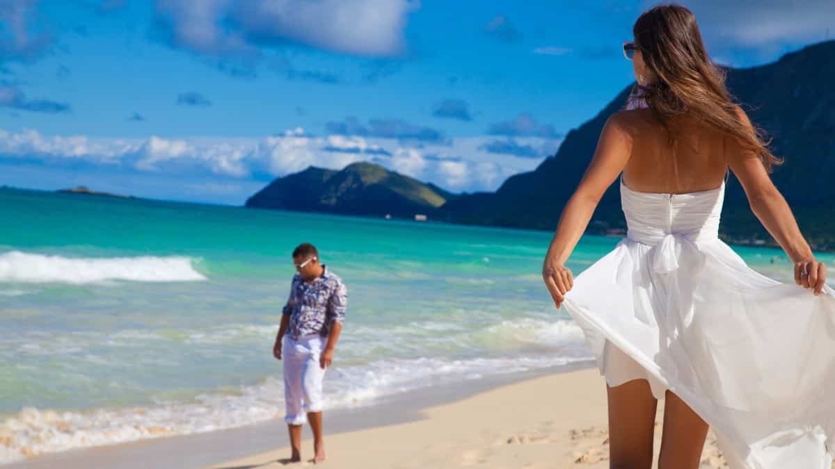 man and woman walking on a beautiful sand beach with lush green mountains in the background