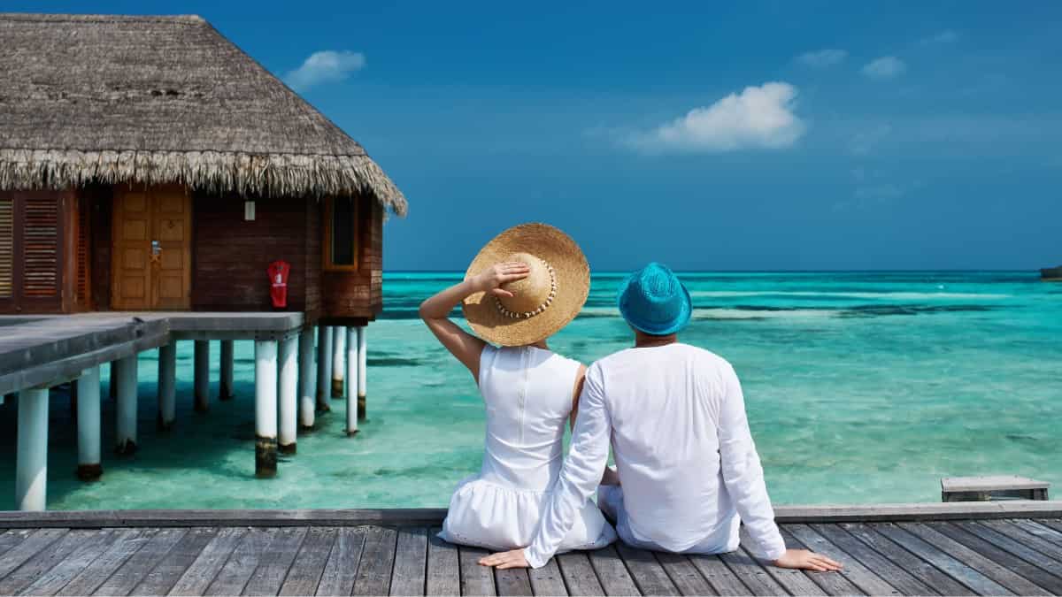 a man and woman sitting on the boardwalk of a Maldives bungalow looking at the turquoise ocean waters