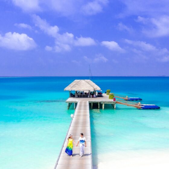 couple walking on the boardwalk of an overwater bungalow with white sandy beach and blue and green waters underneath.