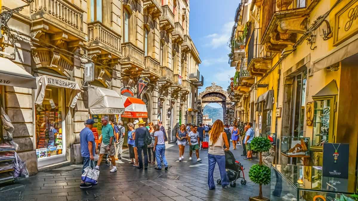 A vibrant shopping street scene from Taormina. Local vendors display their crafts, foods, and traditional Sicilian wares, capturing the essence of Sicilian culture and festivity.